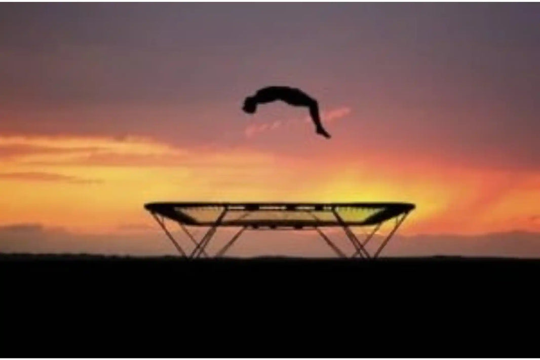 Silhouette of a gymnast performing an aerial flip above a trampoline against a colorful sunset sky.