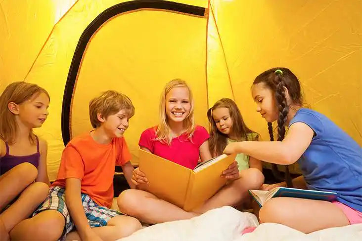 Group of children sitting inside a yellow tent, reading books together during an indoor camping activity.