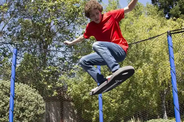 Teen performing a skateboard trick while jumping on a backyard trampoline with a safety enclosure on a sunny day.