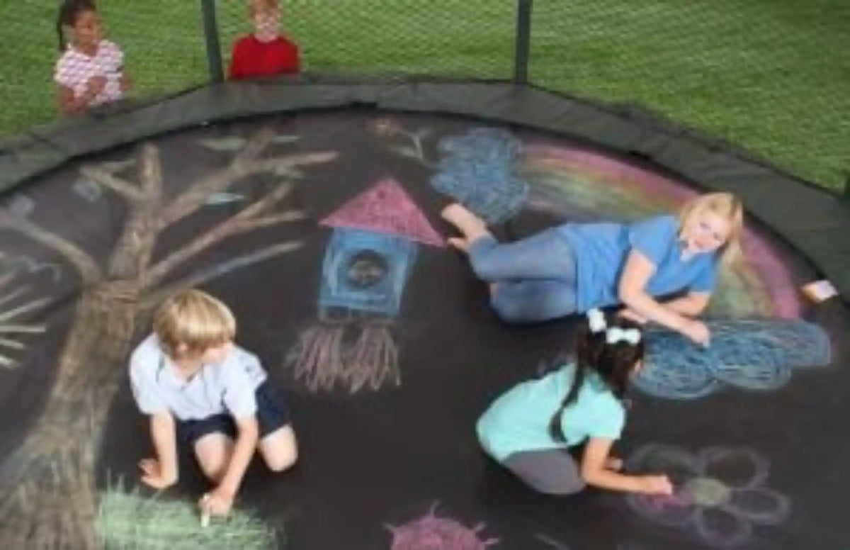 Children drawing colorful chalk artwork on a trampoline mat, including a rainbow, tree, birdhouse, clouds, and flowers, inside a safety enclosure.
